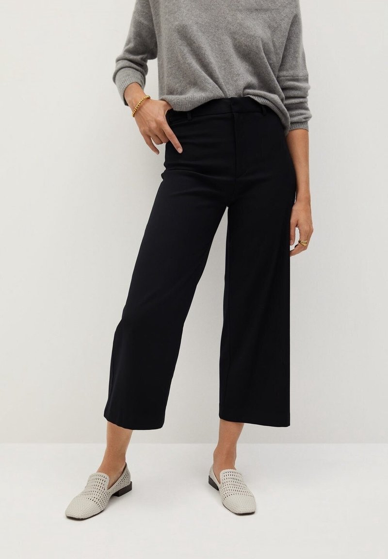 Personne portant un pantalon noir large et coupé court, un pull gris, des chaussures beiges tissées façon slip-on, ainsi qu’un bracelet et des bagues en or, debout devant un fond blanc.