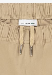 Μπεζ βαμβακερά σορτς με ελαστική μέση, διακοσμημένα με πλεκτά κορδόνια και ετικέτα με το λογότυπο της Lacoste στο κέντρο επάνω μέρος.