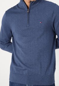 Marineblå zip-up tröja i mjukt tyg, med hög krage, ribbade ärmslut och en diskret logotyp på bröstet.