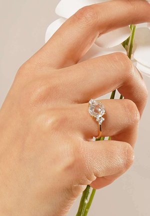 Goldring mit einem zentralen ovalen klaren Edelstein, flankiert von zwei kleineren klaren Steinen. Eine Hand hält einen Stängel mit weißen Blumen im Hintergrund.