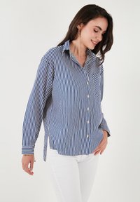 Camicia button-up a righe verticali blu e bianche, con colletto a punta, maniche lunghe e taschino sul petto. Jeans in denim bianco.