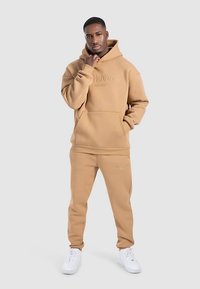 Felpa con cappuccio beige e pantaloni abbinati in tessuto morbido, con tasca frontale e dettaglio con logo. Indossati con scarpe da ginnastica bianche.