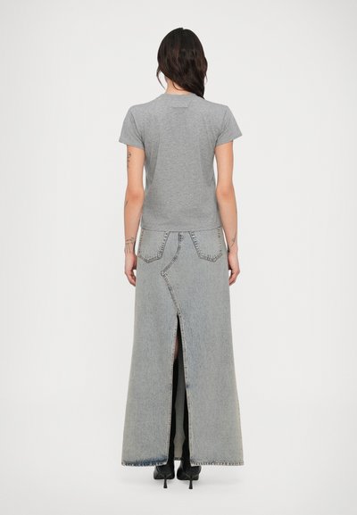 Hosszú sötét hajú nő szürke pólóban, világos farmer hosszú szoknyában hátul sliccel, és fekete magassarkú cipőben, fehér háttér előtt áll.