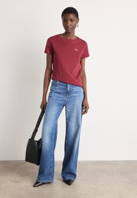 Red cotton t-shirt with short sleeves and a small logo, paired with light blue wide-leg denim jeans. Black pointed-toe heels and a black bag.