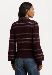 Strikkegenser i burgunder med teksturert kropp, sikksakkmønster og puffede ermer. Brukt over en rutete skjorte, sammen med blå jeans.