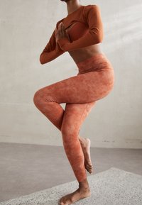 Rust-colored leggings and long-sleeved top featuring a cutout design, textured finish, and smooth fabric, worn in a yoga pose.