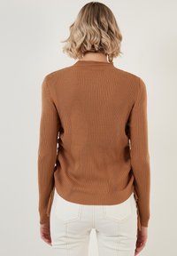 Pruun soonikkudum sweatshirt, millel on keha lähedane disain, pikad varrukad, kõrge kaelus ja külgedel lõhikud, koos valgete pükstega, millel on taga taskud.