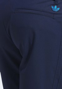 Primeiro plano de tecido azul-marinho mostrando o bolso traseiro e a costura superior com o logotipo trefoil azul da Adidas no canto superior direito.