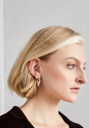 Boucles d'oreilles en argent avec un design incurvé et un petit accent en pierre, portées par un modèle avec des cheveux courts blonds, sur fond neutre.