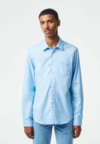 Camicia a maniche lunghe azzurro chiaro con bottoni, realizzata in tessuto morbido, con taschino sul petto, colletto a punta e orlo arrotondato.