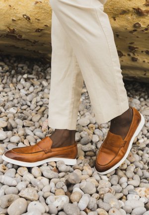 Persona che indossa pantaloni beige corti e mocassini di pelle marrone, in piedi su ciottoli grigi lisci vicino a una grande roccia gialla.