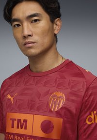 Crvena nogometna majica s teksturiranim uzorkom, narančasti Puma logo i narančasti grb Valencia CF na prsima. Ojačana logotipima sponzora.