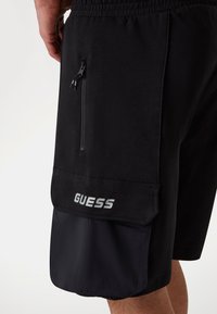 Schwarze Shorts mit elastischem Bund und Reißverschlusstasche, aus einem strukturierten Stoff und mit dem "GUESS"-Logo in Silber am unteren Bein.