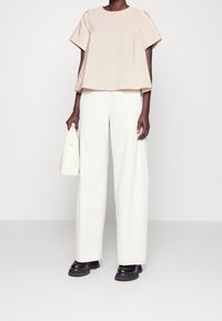 Beige lyhythihainen toppi, leveälahkeiset valkoiset housut ja mustat paksupohjaiset kengät. Malli pitää kädessään pientä valkoista laukkua. Sileä kangas, moderni leikkaus.