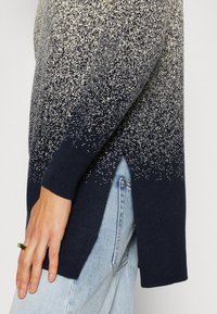 Vue latérale d'une personne portant un pull bleu et blanc moucheté avec une fente sur le côté, un jean bleu clair et une bague verte à son doigt.