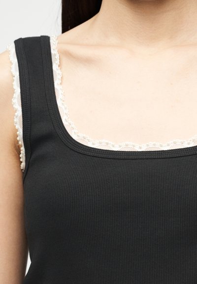 Femme portant un débardeur noir côtelé superposé à un caraco blanc orné de dentelle, dévoilant le clavicule et la zone de l'épaule.