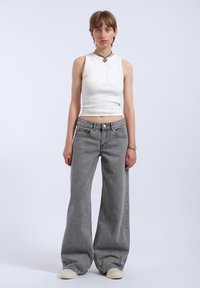 Un haut blanc sans manches ajusté associé à un jean gris taille haute et à pattes d'éléphant, accompagné de baskets beiges. Les accessoires comprennent des colliers superposés.