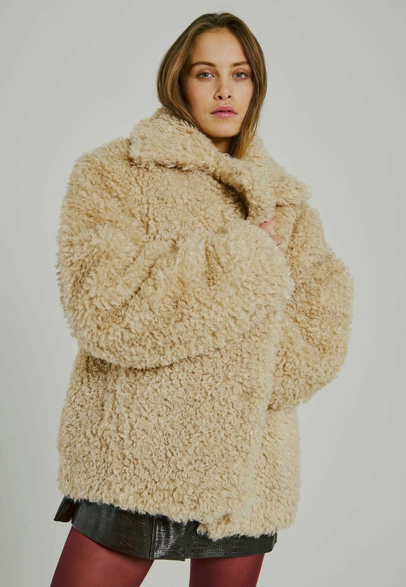 Cappotto beige, peloso e oversize realizzato in tessuto peluche con ampio colletto e spalle cadenti, progettato per il calore e il comfort.