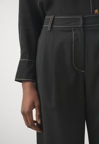 Černé kalhoty na míru s bílými švy a širokým páskem. Obsahují přední kapsu a hladký textilní povrch. Ruka položená na pase.