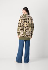 Adīts kardigans ar ģeometrisku rakstu smilšu, zaļā un brūnā krāsā, ar garu, brīvu piegriezumu un ribotām aprocēm. Apvienots ar platām biksēm.