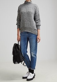 Grå stickad turtleneck-tröja, lös passform; blå slitna jeans; svart fransad ryggsäck; vita snörskor med tjocka svarta sulor.