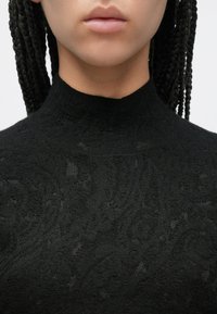 Μαύρο υφασμάτινο τοπ με ψηλό γιακά, διακοσμημένο με μια διακριτική παγιέτα, κατασκευασμένο από μαλακό ύφασμα, με εφαρμοστή σιλουέτα και ψηλό λαιμό.