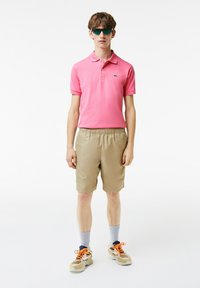 Rosa pikétröja med korta ärmar, beigefärgade shorts, grå strumpor och bruna sneakers med orangea accenter. Modellen bär gröna solglasögon.