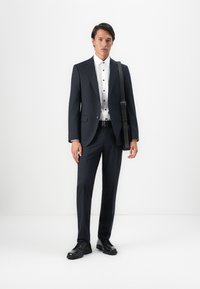 Man wearing dark navy suit, white shirt with black buttons, black shoes, and carrying black shoulder bag, standing against white background.