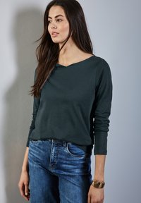 Tummanvihreä pitkähiha toppi, jossa on V-kaula-aukko, yhdistettynä vaaleansinisiin farkkuihin. Kangas näyttää pehmeältä ja siinä on yksinkertainen siluetti.