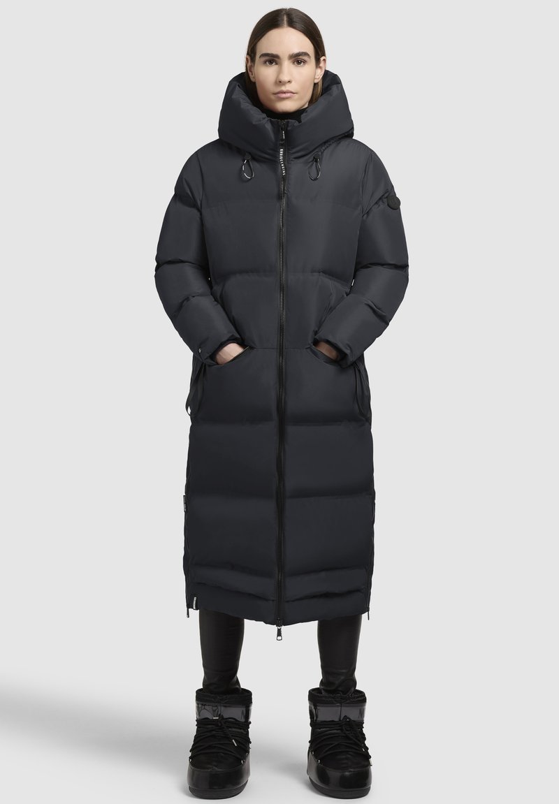 Abrigo de plumas largo negro con cuello alto, cierre de cremallera y bolsillos laterales. Tiene un diseño acolchado y una tela texturizada. Llevado con botas negras.