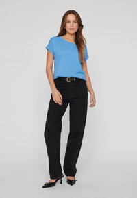 Femme debout portant une chemise bleue à manches courtes, un pantalon taille haute noir avec une ceinture, et des chaussures à talons noires sur un fond uni.