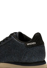 Baskets à paillettes noires avec un onglet arrière texturé, portant le nom de la marque "WODEN". La semelle est de couleur claire avec une finition lisse.