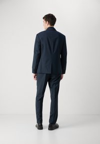 Completo blu navy con una vestibilità su misura, apertura singola sul retro e polsini con bottoni. Il materiale sembra liscio con una leggera lucentezza. Scarpe nere incluse.