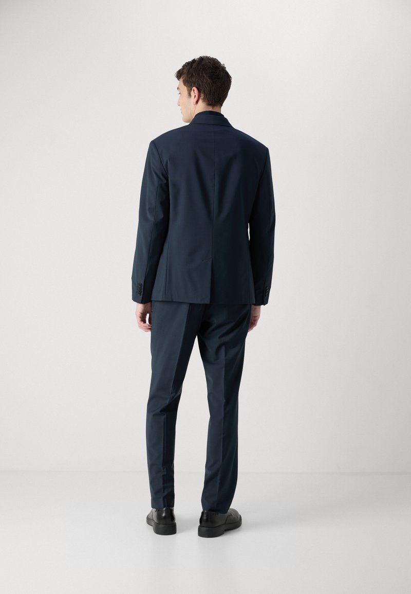Costume marin avec une coupe ajustée, une fente simple à l'arrière et des poignets boutonnés. Le matériau semble lisse avec un léger brillantage. Chaussures noires incluses.