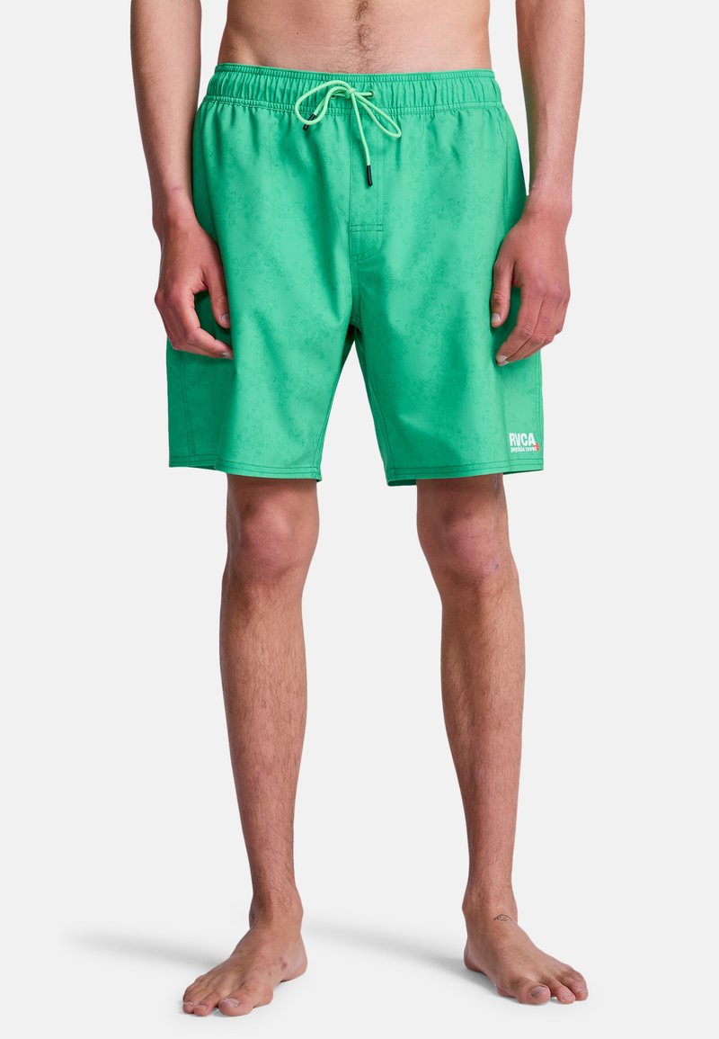 Homme portant un short de bain vert à taille élastique avec cordon de serrage, debout pieds nus devant un fond uni.