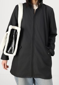 Cappotto impermeabile nero con zip frontale, maniche lunghe e due tasche laterali. Abbinato a una borsa bianca e nera con texture e tracolla pelosa.