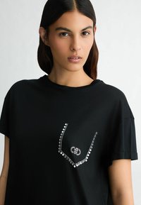 Must have lühikeste varrukatega must t-särk, valmistatud pehmest kangast, millel on rinnal dekoratiivne kristallidest kujundus "V" kuju. 