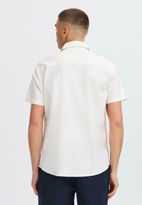 Camisa de manga corta de color claro, hecha de una tela de textura suave, con cuello tipo punta, dobladillo recto y sin patrones visibles.
