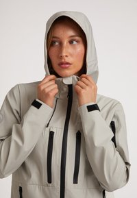 Lys grå vandtæt jakke med hætte, med sorte lynlåse og velcro-detaljer, designet med en strømlinet pasform og praktiske lommer.