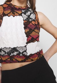 Neulecroppitoppi, jossa on valkoinen keskikohta värikkäillä applikaatiokuvioilla oranssissa, punaisessa ja mustassa. Korkea kaulus ja teksturoitu kangas.