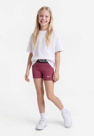 Smilende pige med langt blondt hår iført hvid oversized t-shirt, vinrøde Nike-shorts, hvide sokker og hvide sneakers, stående på hvid baggrund.