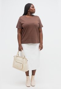 Γυναίκα με πλεξούδες που φοράει καφέ πουκάμισο και λευκή φούστα, κρατώντας μπεζ τσάντα, κοιτάζοντας στο πλάι σε λευκό φόντο.