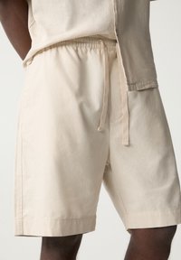 Shorts beige en mélange de coton avec une ceinture élastique et un cordon de serrage. Comprennent des poches latérales et une coupe décontractée. Texture lisse, longueur au genou.