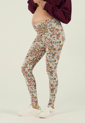 Personne enceinte portant un legging de maternité à motifs floraux et des baskets blanches, soulevant un haut bordeaux pour dévoiler son ventre.