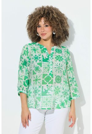 Vrouw met krullend haar, die een groen-wit gebloemd blouse met driekwartmouwen en een witte broek draagt, staand tegen een witte achtergrond.