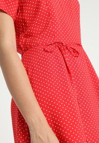Robe rouge à pois blancs avec manches courtes, présentant une taille cintrée et un détail noué. Le tissu est lisse avec des pois blancs répartis uniformément.