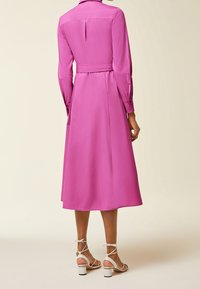 Lungo vestito rosa con vita aderente, gonna fluttuante e maniche con polsini bottonati. Paio di décolleté bianche con cinturini e tacco a blocco visibile nella parte inferiore.