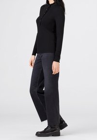 Femme debout portant un top noir à manches longues avec des détails de boutons, un jean large sombre avec des ourlets bruts et des bottes noires à lacets.