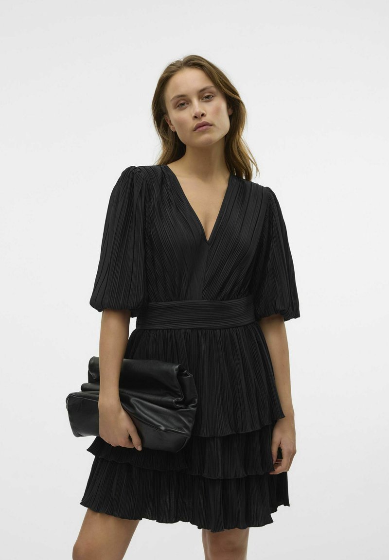 Czarna plisowana sukienka z głębokim dekoltem w serek, krótkimi bufkami i warstwowym dołem. Połączona z wgniecioną czarną torebką kopertową.