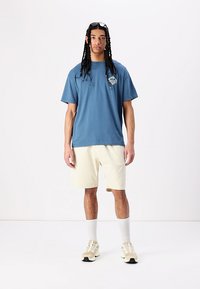 T-shirt en coton bleu avec un design graphique sur la poitrine, associé à un short beige clair. Le modèle porte des chaussettes blanches et des baskets crème.